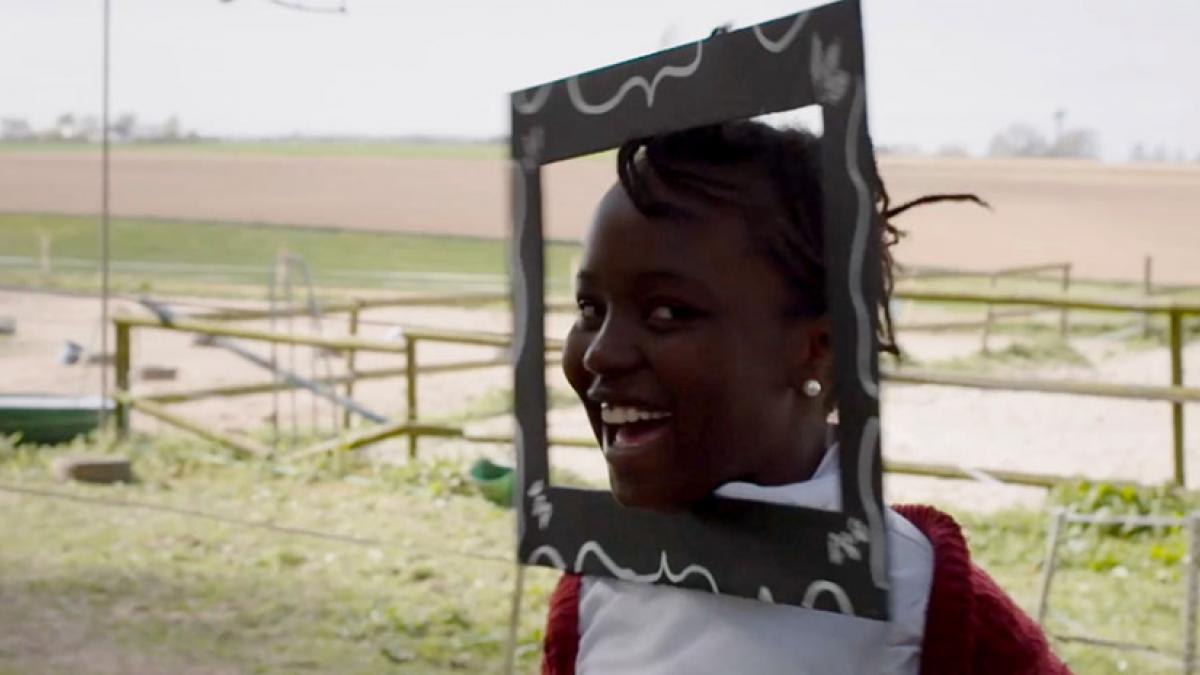 A laughing girl with a picture frame attached to her face, with fenced agricultural fields in the background.