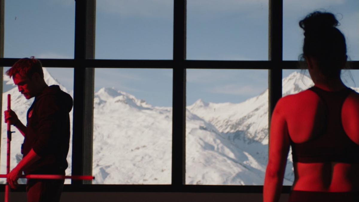 In the foreground, a woman in a sport tank with her hair in a bun, her back to the camera. In the middle ground, a bearded man in a hoodie with his back to her. In the background, windows looking out over snowy mountains.