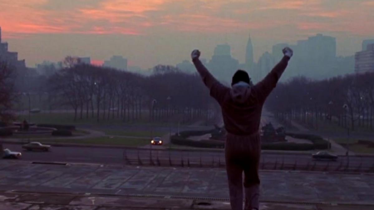 Wide shot of a man in sweatpants and hoodie, with his back to the viewer, holding his arms high in the air. In the background is a Philedelphia citscape at sunrise.