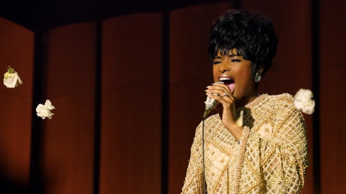 A medium shot of a Black woman in a vintage yellow gown and retro hairstyle singing into a microphone.