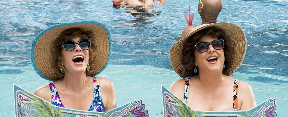 Two middle-aged women with delighted expressions, wearing swimsuits, sunhats, and sunglasses. They are floating chest-deep in a swimming pool while holding menus.