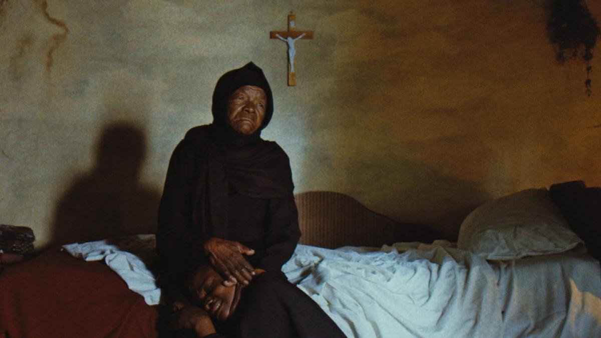 An elderly Black woman in long black robes and headscarf sits on a bed, cradling a younger Black man's head in her lap.