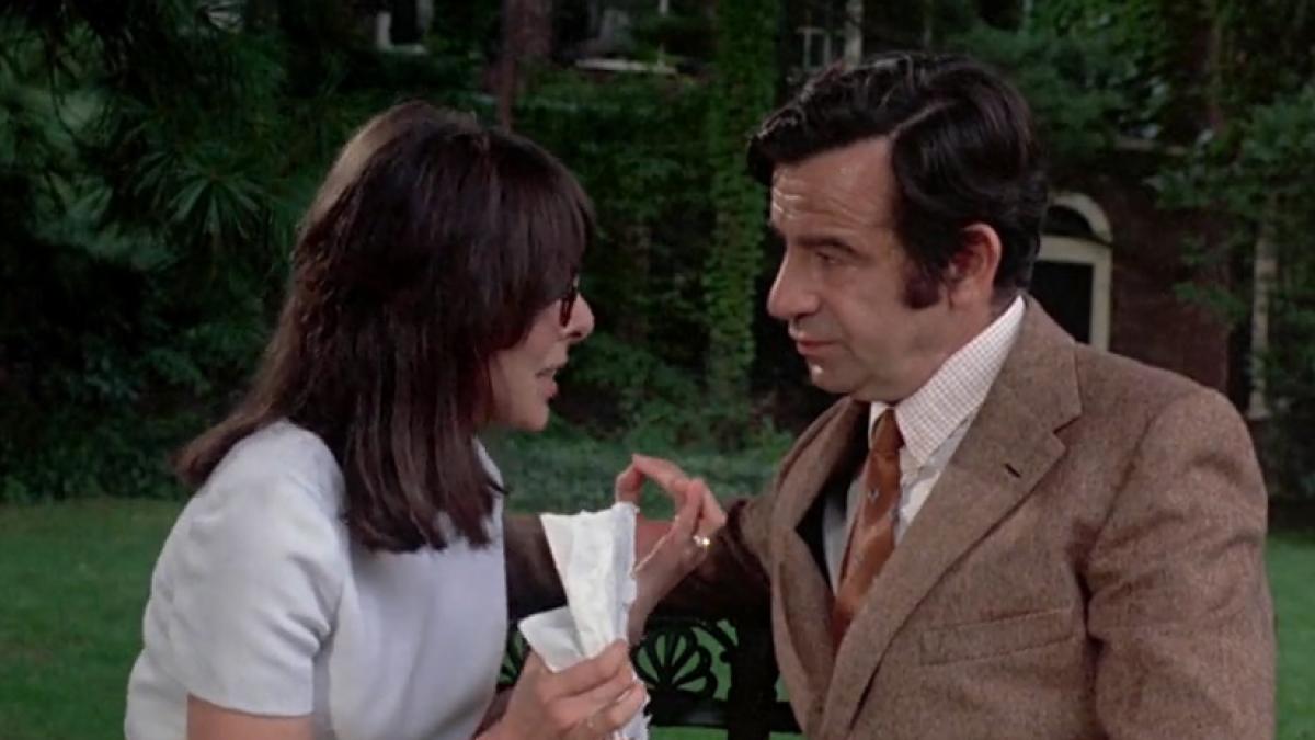 A woman in a white blouse and a man in a brown suit sit together, talking urgently, in a large, grassy outdoor area.
