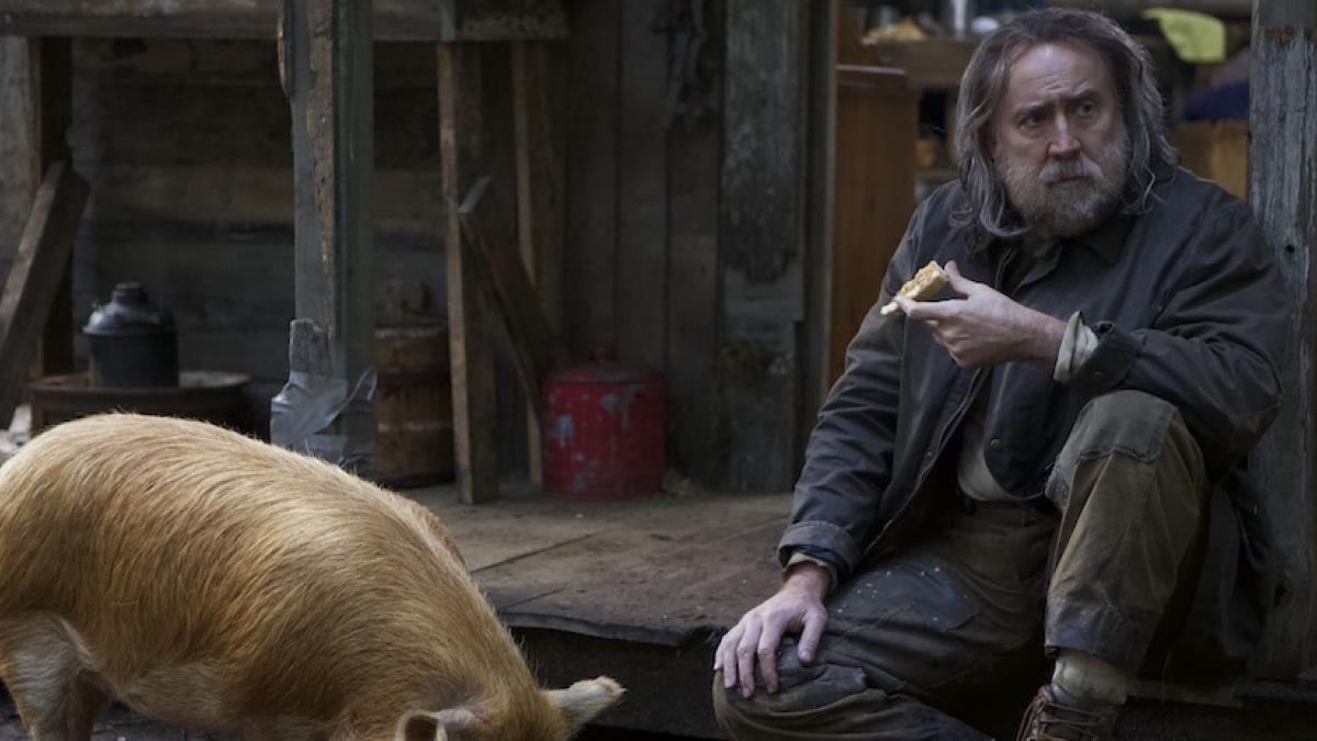 A man in outdoor wear sits in front of a cabin in the forest while a pig eats from a bowl in front of him.