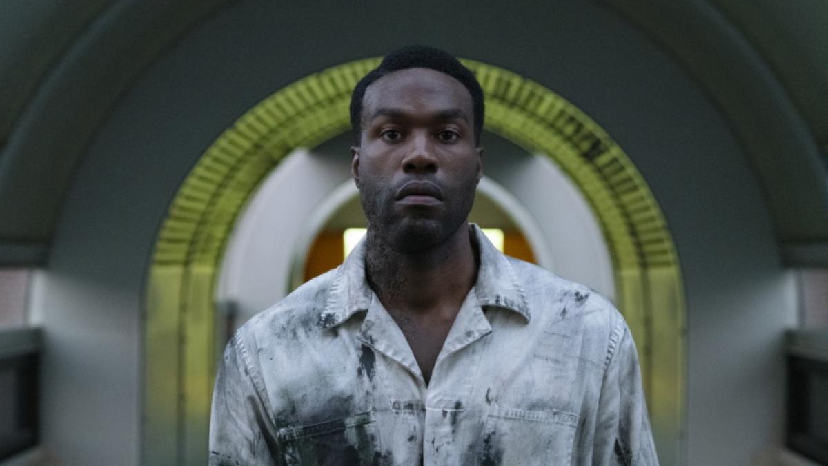 Medium shot of a Black man standing in an arched hallway, facing the camera, his white shirt smeared with black paint.