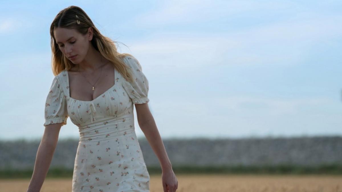 A medium shot of a young woman in a white summer dress walking through a wheat field.