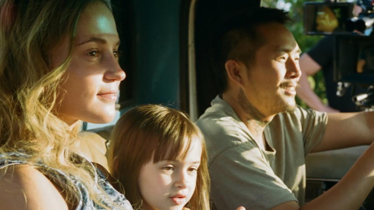 Medium shot of (left to right) a woman, little girl, and man sitting in the front scene of a truck, passing by sunny vegetaion.