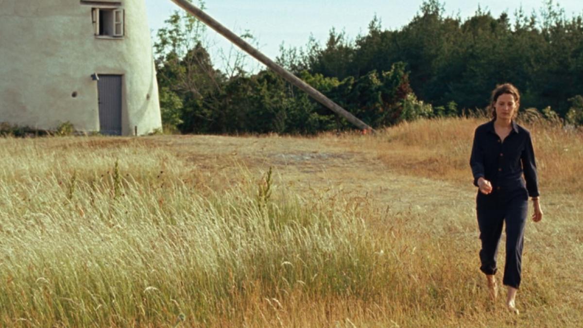 Wide shot of a woman in dark clothes walking through a dry brown field, with a windmill partially visible in the background.
