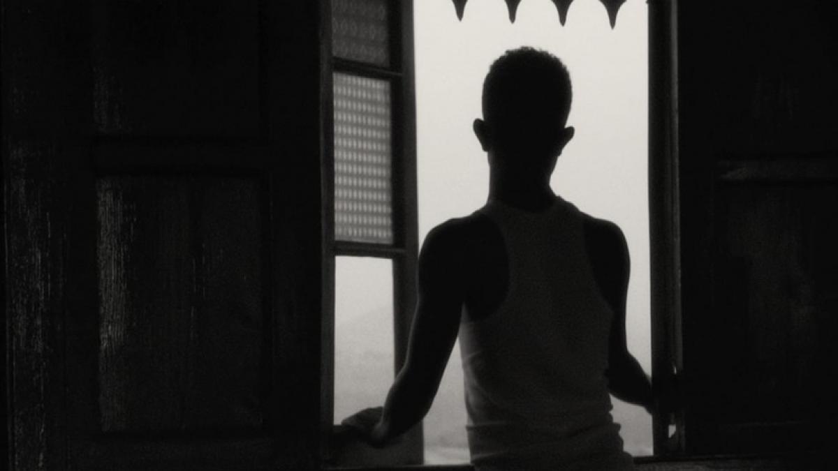 Medium black-and-white shot from behind of an adolescent boy standing in front of an open window.
