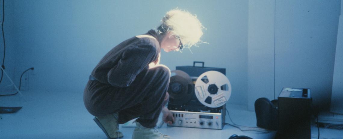A woman crouches in profile, inspecting a real-to-real audio device.