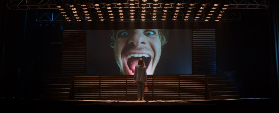 A woman standing in front of a giant screen, which is showing an image of a man's laughing face.