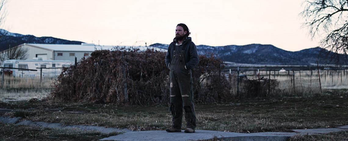 A bearded man in a hoodie and coveralls standing in a vacant lot in front on an overgrown fence, with mountains in the distance.