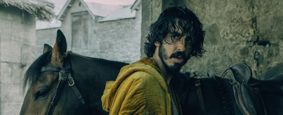 Medium-close shot of a bearded man with dark dishelved hair and a yellow cloak, standing in front of a dark horse.