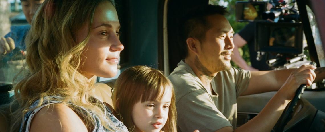 Medium shot of (left to right) a woman, little girl, and man sitting in the front scene of a truck, passing by sunny vegetaion.