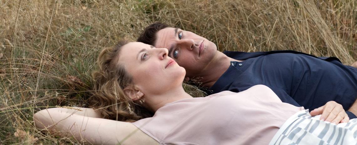 Medium shot of a middle-aged woman laying in the grass with a slightly younger man; she is looking up into the sky, he is looking at her.