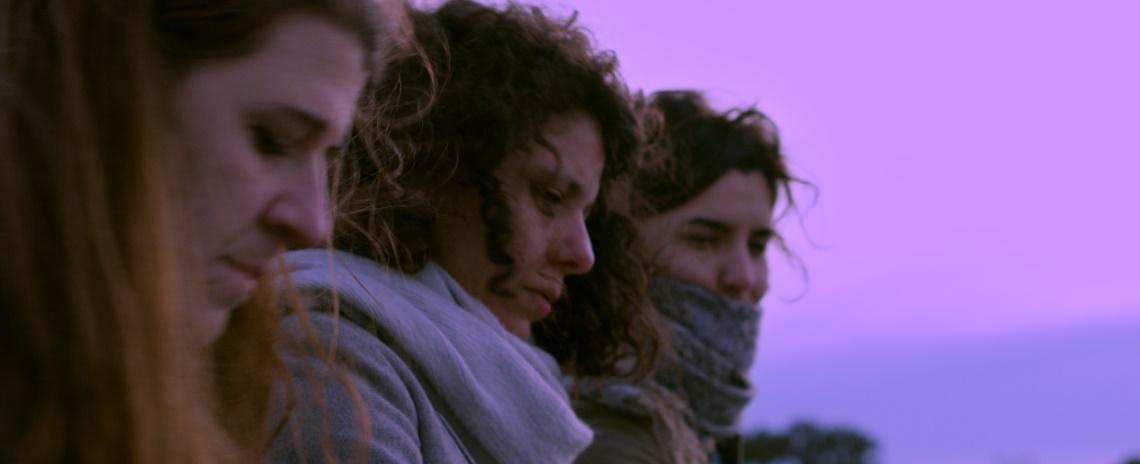 Medium-close profile shot of three women arranged in a row, facing right. In the background is a pink sky.