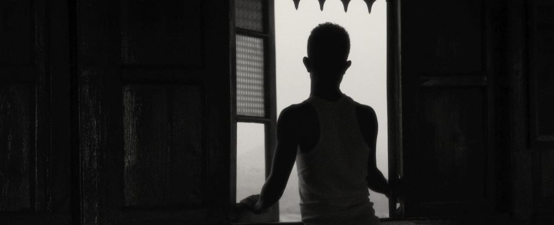 Medium black-and-white shot from behind of an adolescent boy standing in front of an open window.