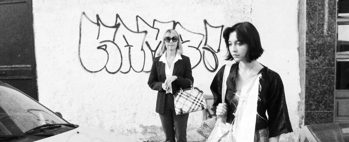 Black and white medium shot of two stylishly-dressed women starting to cross a street in front of a wall with graffiti.