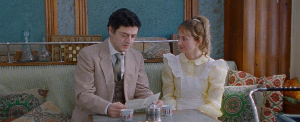 Medium shot of a man in a suit and a woman in a frilly dress sitting in a cafe.